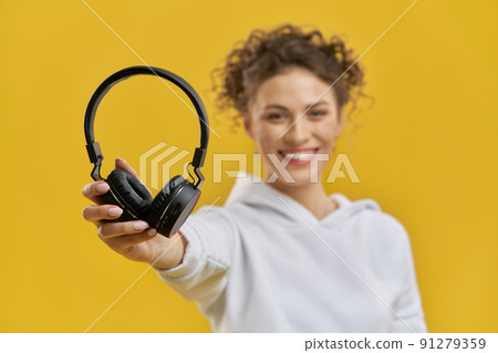 Neat female hand holding up black headphones indoor. Close up view of earphones supported by girl's hand, with blurred figure of smiling woman, isolated on orange studio background. Concept of music. 91279359