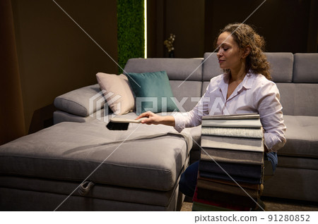 Pretty young woman- sales manager, in furniture store, holding upholstery fabric samples in her hands and cleaning sofa and pillows with a dustpan in home design studio Pretty young woman- sales manager, in furniture store, holding upholstery fabric samples in her hands and cleaning sofa and pillows with a dustpan in home design studio 91280852