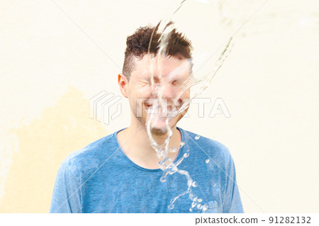 Wet smiling young caucasian man in blue t-shirt with closed eyes in water splash, jet of water on a yellow background. Stress, shock. Front view, portrait 91282132