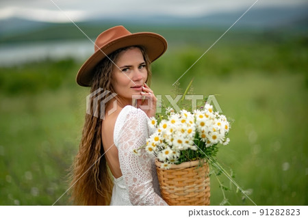 A middle-aged woman in a white dress and brown hat stands on a green field and holds a basket in her hands with a large bouquet of daisies. In the background there are mountains and a lake. A middle-aged woman in a white dress and brown hat stands on a green field and holds a basket in her hands with a large bouquet of daisies. In the background there are mountains and a lake. 91282823