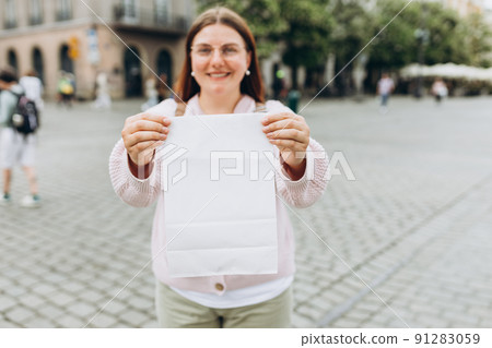 Young happy redhead woman in glasses holding take away paper bag over street background. White Packaging template mock up. Delivery service concept. Blank snack white paper bag 91283059