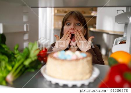 Young woman looks in surprise at an appetizing cake in the refrigerator Young woman looks in surprise at an appetizing cake in the refrigerator 91286378