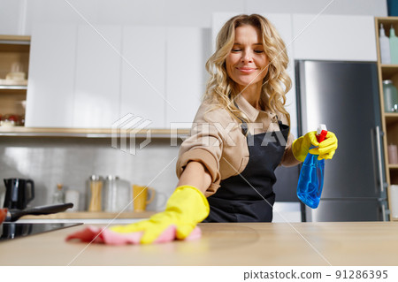 Pleasant middle aged woman cleaning countertop in kitchen with rag and cleaning disinfectant agent Pleasant middle aged woman cleaning countertop in kitchen with rag and cleaning disinfectant agent 91286395