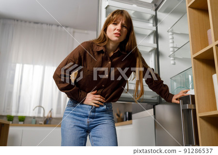 Young woman near empty refrigerator feels pain in her stomach. Hungry girl holding on to her abdomen Young woman near empty refrigerator feels pain in her stomach. Hungry girl holding on to her abdomen 91286537