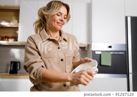 Happy housewife woman wiping plate with dry towel in kitchen after dish washing Happy housewife woman wiping plate with dry towel in kitchen after dish washing 91287120