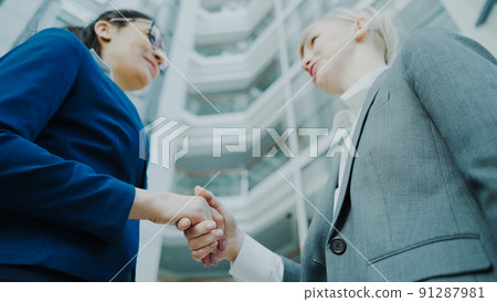 Low angle shot of businesswoman shaking hands and talking with female business colleague in suit in hall of modern office building 91287981