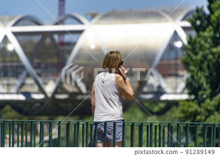Woman. Person. Middle-aged lady talking on the phone in a park in Madrid. Metal bridge of the Madrid River park in the background. Clear day, in Spain. Europe. Horizontal photography. 91289149