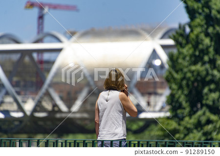 Woman. Person. Middle-aged lady talking on the phone in a park in Madrid. Metal bridge of the Madrid River park in the background. Clear day, in Spain. Europe. Horizontal photography. Woman. Person. Middle-aged lady talking on the phone in a park in Madrid. Metal bridge of the Madrid River park in the background. Clear day, in Spain. Europe. Horizontal photography. 91289150