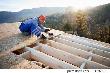 Carpenter hammering nail into OSB panel on the roof top of future cottage in the evening. Man worker building wooden frame house. Carpentry and construction concept. 91292586