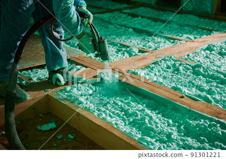 Male builder insulating wooden frame house. Cropped view of man worker spraying polyurethane foam on floor inside of future cottage, using plural component gun. Construction and insulation concept. 91301221