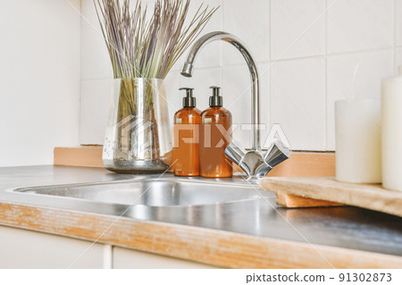 A fragment of the interior of a light home kitchen with beige furniture in a minimalist style with a sink, mixer tap, soap and candles 91302873