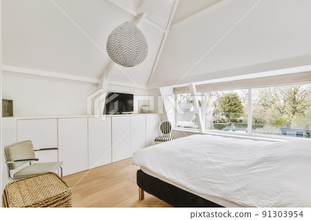 Modern interior design of a small attic bedroom with white walls and a soft cozy bed, storage places, a panoramic window and a TV in front of the bedamic window 91303954
