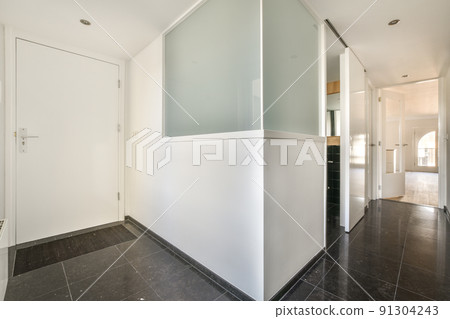 Narrow corridor with white walls and doors leading to spacious room with windows and parquet floor in modern apartment 91304243