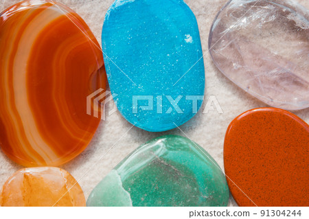 Background of semi-precious multicolored polished stones, pebbles on light white sand on a seashore beach. Stones macro shot. Mineral rocks backdrop wallpaper. Precious jasper stone. Geology concept. 91304244