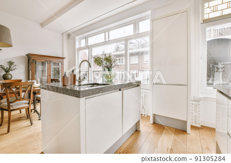 Modern minimalist style interior design of studio apartment with open white kitchen and dining zone with table and chairs illuminated by loft style lamp 91305284