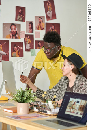 Retoucher sitting at table and consulting with photo editor about images on computer, they working in team at office 91306340