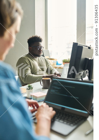 African young man sitting at table in front of computer monitor and typing with his colleague sitting opposite him and working on laptop 91306435