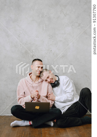 Young gay couple sitting on floor using laptop, using headphones listen to music together, hugging or embracing. 91307760