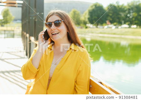 Smiling beautiful woman talking on phone on river background. Happy woman with yellow shopping bag is using a smartphone outdoors, summer time. Traveler. Women posed against nature park Smiling beautiful woman talking on phone on river background. Happy woman with yellow shopping bag is using a smartphone outdoors, summer time. Traveler. Women posed against nature park 91308481