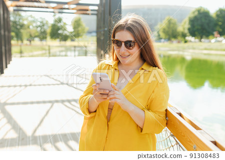 Beautiful stylish woman walking in the park on summer day and holding mobile phone. Phone Communication. Urban lifestyle concept. Traveler Beautiful stylish woman walking in the park on summer day and holding mobile phone. Phone Communication. Urban lifestyle concept. Traveler 91308483