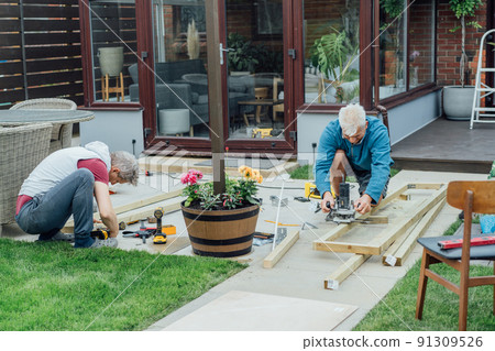 Adult son and father carpenter working together, handle wooden timbers in the garden. Patio construction by your own. DIY, Do it yourself. Home renovation, improvement, refurbishment. Selective focus Adult son and father carpenter working together, handle wooden timbers in the garden. Patio construction by your own. DIY, Do it yourself. Home renovation, improvement, refurbishment. Selective focus 91309526
