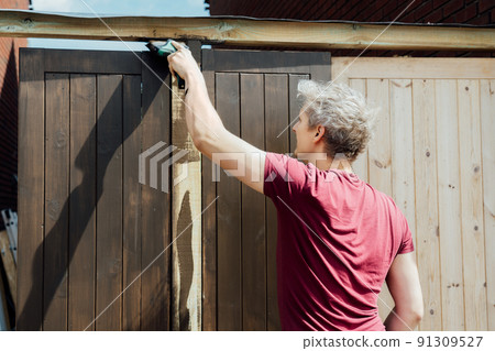 Young man worker paints with a brush a wooden board fence gate. DIY, Do it yourself concept. House improvement. Home renovation and refurbishment. Selective focus, copy space. Young man worker paints with a brush a wooden board fence gate. DIY, Do it yourself concept. House improvement. Home renovation and refurbishment. Selective focus, copy space. 91309527