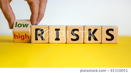 Hand is turning a cube and changes the words 'high risks' to 'low risks' or vice versa. Beautiful yellow table, white background, copy space. Business concept. Hand is turning a cube and changes the words 'high risks' to 'low risks' or vice versa. Beautiful yellow table, white background, copy space. Business concept. 91310051