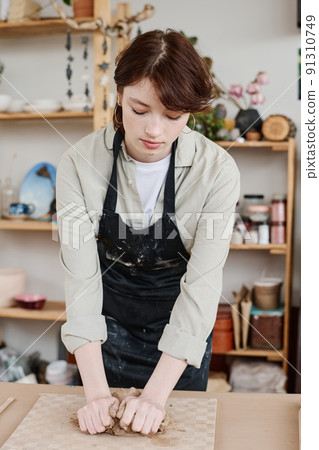 Young skilled female potter in apron bending over table while kneading clay 91310749