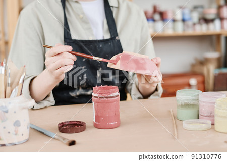 Young creative woman in apron holding paintbrush and clay bowl over table 91310776