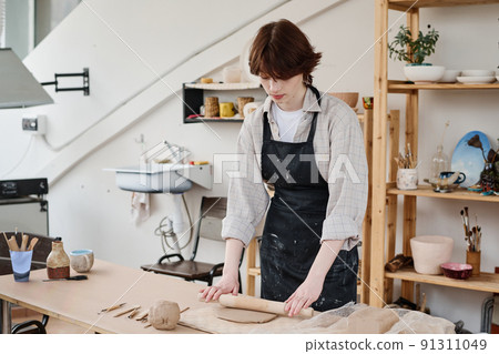 Young woman in black apron flattening piece of clay with rolling pin in workshop Young woman in black apron flattening piece of clay with rolling pin in workshop 91311049