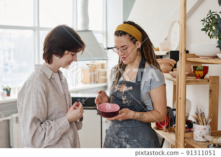 Young female clerk of handmade earthenware shop showing clay bowl to buyer Young female clerk of handmade earthenware shop showing clay bowl to buyer 91311051