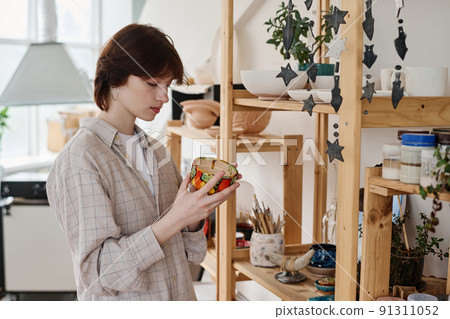 Young brunette woman choosing gift in earthenware shop while holding clay bowl Young brunette woman choosing gift in earthenware shop while holding clay bowl 91311052