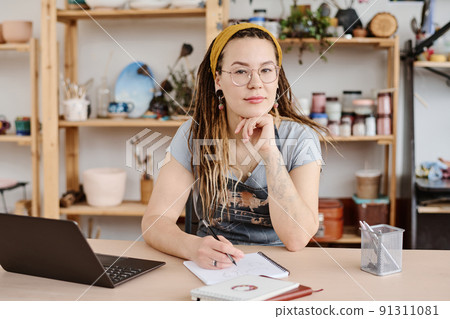 Young modern female in casualwear and eyeglasses sitting by workplace 91311081