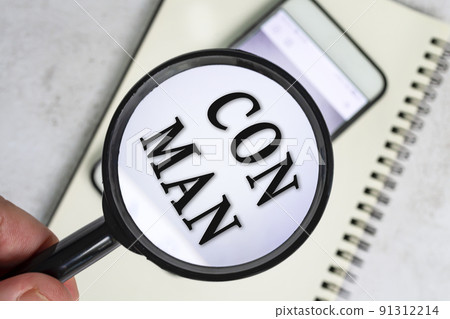 A man's hand holds a black magnifying glass by the handle with con man text. 91312214