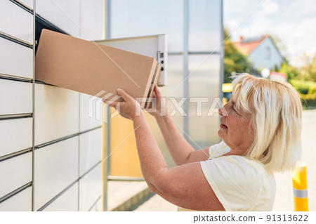 50s caucasian woman getting parcel from cell of automatic post terminal outdoors. 50s caucasian woman getting parcel from cell of automatic post terminal outdoors. 91313622