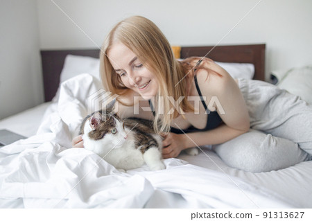 Portrait of young blonde 30s woman holding unhappy tabby cat with green eyes. Female hugging cat in hands at morning bed Portrait of young blonde 30s woman holding unhappy tabby cat with green eyes. Female hugging cat in hands at morning bed 91313627