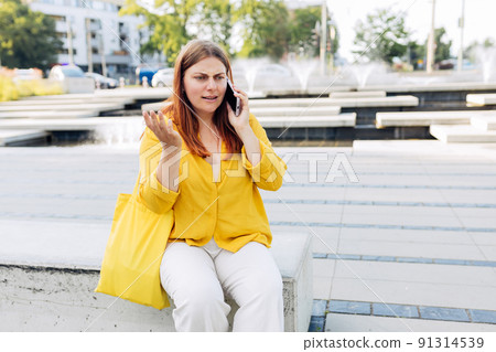 Upset young woman talking over phone disappointed hear bad news, sad pensive girl speaking having unpleasant smartphone conversation and siting on bench on city street. Negative emotions 91314539