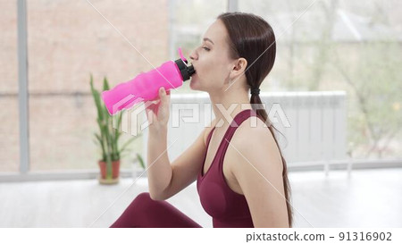 a young woman in a burgundy tracksuit drinks water on a mat in a gym. 91316902