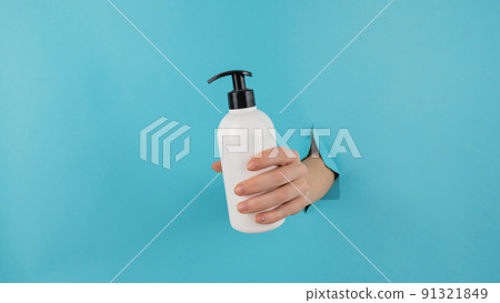A woman's hand sticks out of a hole in a blue paper background and holds a skin lotion pump. 91321849