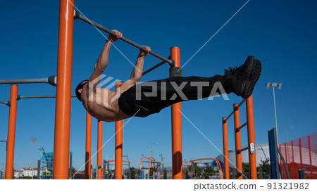 Shirtless man doing blanche on horizontal bar outdoors.  91321982