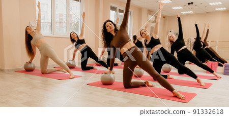 Group of middle aged women with fitness instructor in sportswear leggings and tops, doing Pilates with the ball on the yoga mat near the large mirror. Female fitness yoga routine concept Group of middle aged women with fitness instructor in sportswear leggings and tops, doing Pilates with the ball on the yoga mat near the large mirror. Female fitness yoga routine concept 91332618