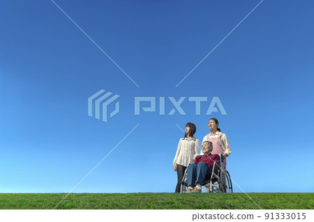 Two women caring for an elderly woman in a wheelchair against the backdrop of the blue sky. Image of family, old age, long-term care 91333015