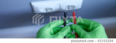 Gloved hands are holding a digital multimeter near the socket Gloved hands are holding a digital multimeter near the socket 91333204