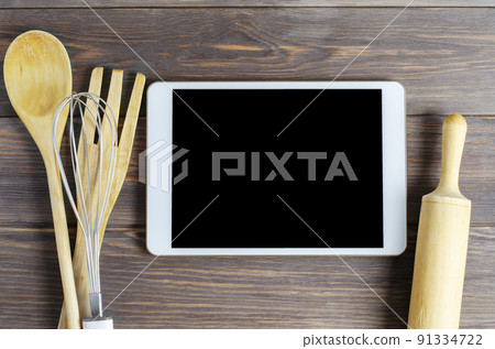A tablet computer and cooking battery on a brown wooden background. Copy space. Concept of cooking recipes A tablet computer and cooking battery on a brown wooden background. Copy space. Concept of cooking recipes 91334722