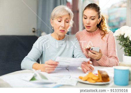 Elderly woman with adult daughter using smartphone and checking utility bills 91335016