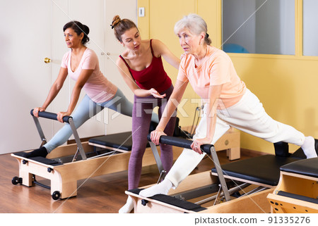 Young pilates instructor assisting elderly woman to do exercises on reformer 91335276