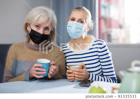 Mature mother and her adult daughter in masks are sitting at a table, holding mugs of tea 91335531