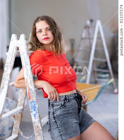 Playful woman in red t-shirt and denim shorts posing in refurbished room Playful woman in red t-shirt and denim shorts posing in refurbished room 91335574