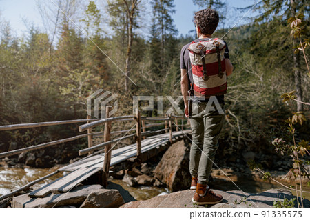 Rear on man with backpack standing on rocks in mountains at wooden bridge over river. 91335575