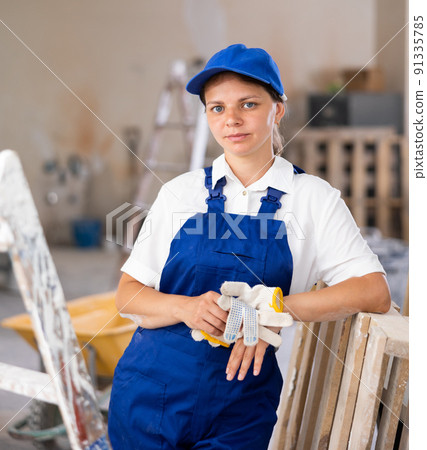 Portrait of contented young female builder in blue overalls during the repair of cottage Portrait of contented young female builder in blue overalls during the repair of cottage 91335785